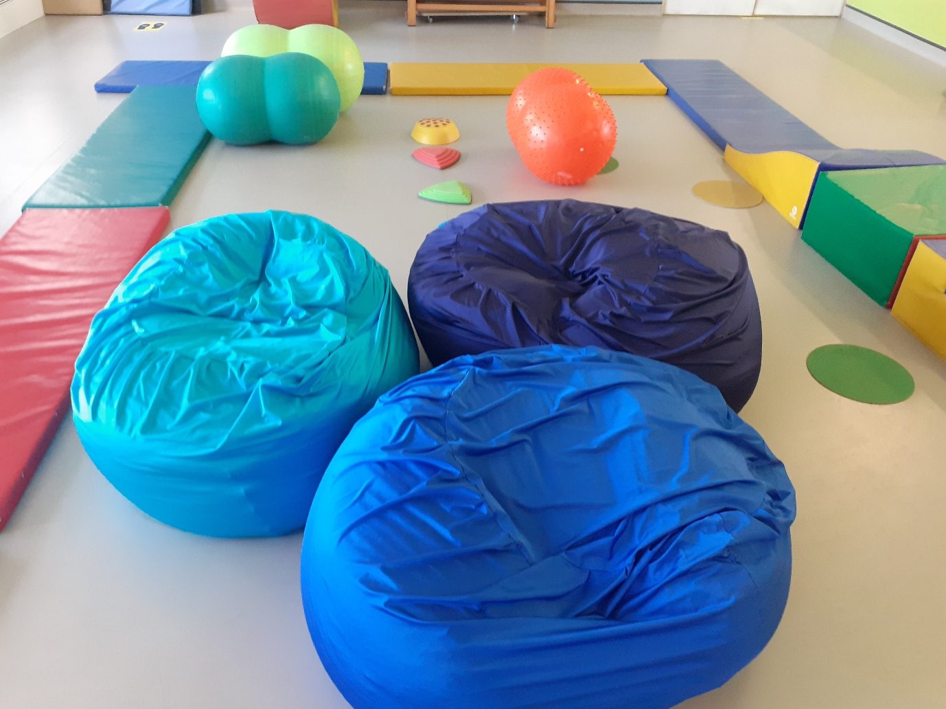 Three shiny stretch-lycra sensory beanbags in turquoise, navy and royal blue on the floor of a sensory room, with gym mats and therapy balls behind