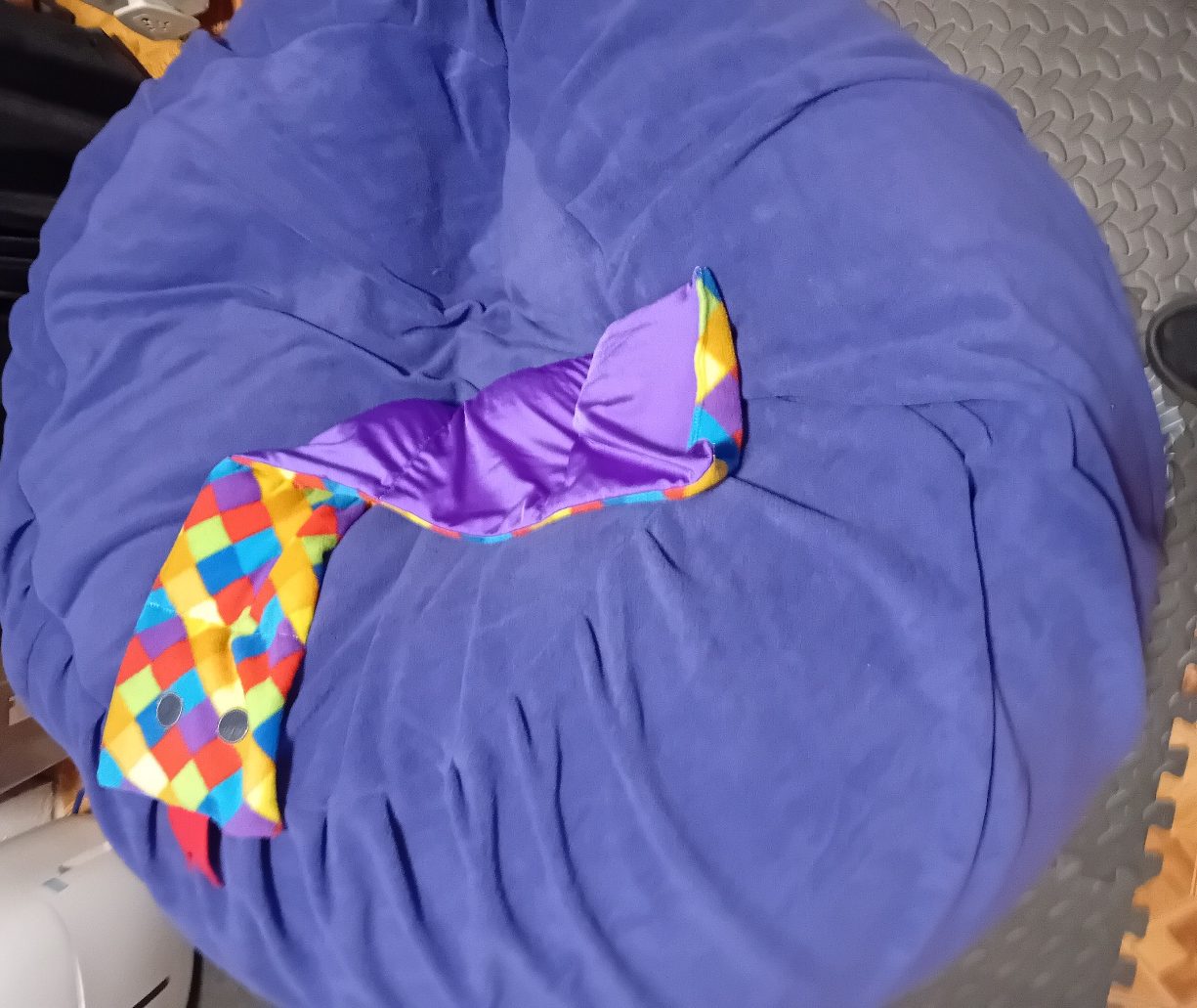 A rainbow-check weighted snake with button eyes and a red tongue, lying on a purple fleece sensory beanbag, with its shiny purple underside showing