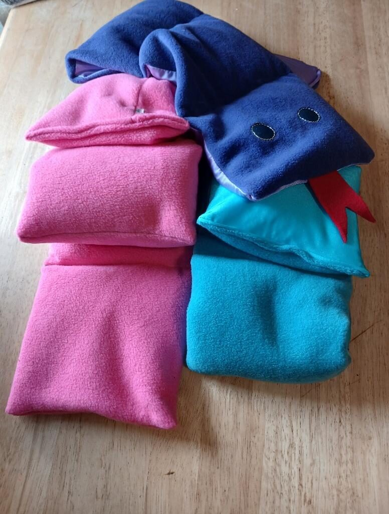 A long pink fleece weighted lap pad and a turquoise lycra-and-fleece one, folded on a wooden table, with a navy weighted snake with button eyes and a red tongue lying across them