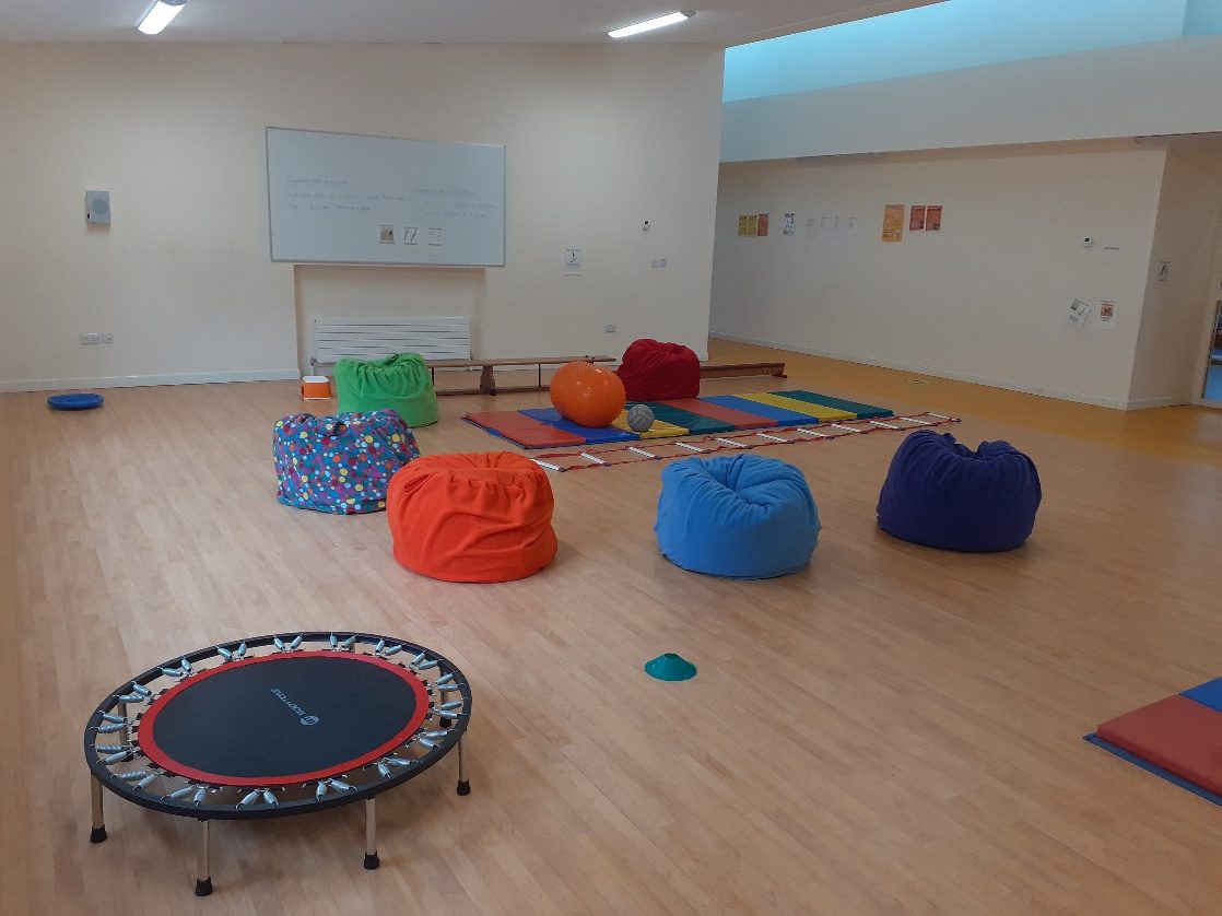 A school sensory room with five fleece beanbags in green, spotted blue, orange, sky blue and purple, a mini trampoline and a row of gym mats